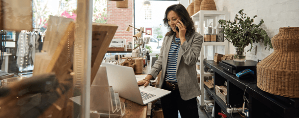 A woman stands in front of a laptop talking on her smartphone.