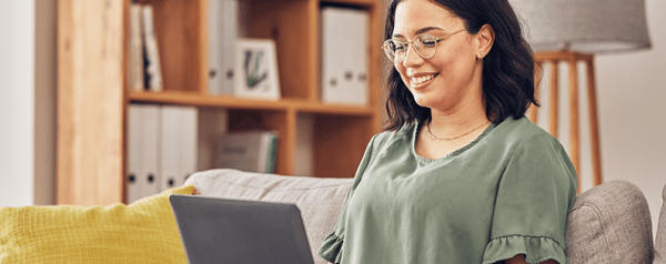 A woman with glasses sits on a sofa with a laptop.
