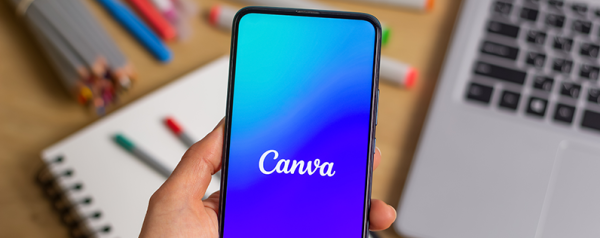 A hand holding a smartphone displaying the Canva logo on screen, plus a laptop and a notebook in the background