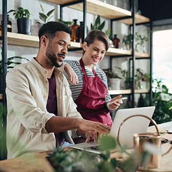Two people stand in front of a laptop with shelves of plants in the background.