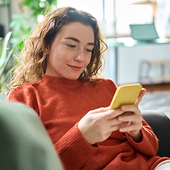 A woman in an orange sweater holds a smartphone and looks at the screen.