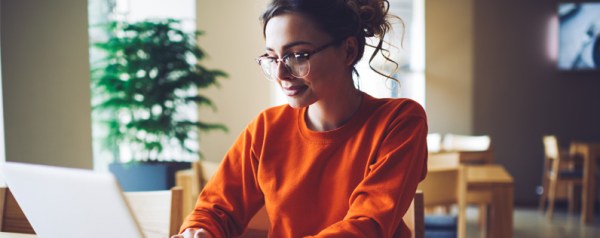 A young woman wearing glasses and an orange sweater sitting in a cafe or office setting while working on a laptop.