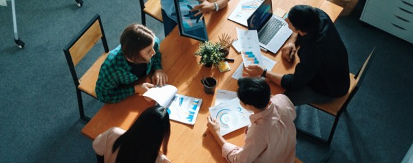 A group of people with laptops sit around a conference table with papers spread out.