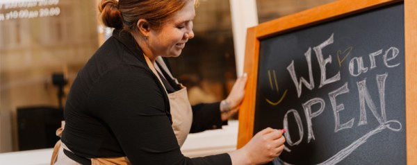 A woman in an apron writes "WE ARE OPEN" on a chalkboard.