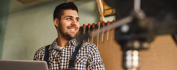 A smiling male tradesperson in a workshop using a laptop.
