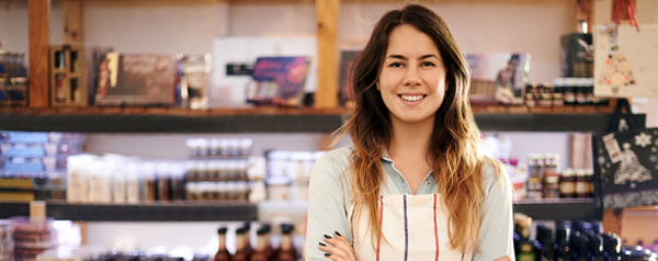 A smiling female small business owner standing in her store with her arms crossed, wearing a striped apron.