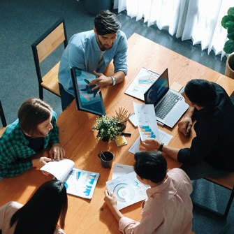 A group of people with laptops sit around a conference table with papers spread out.
