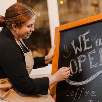 A woman in an apron writes "WE ARE OPEN" on a chalkboard.