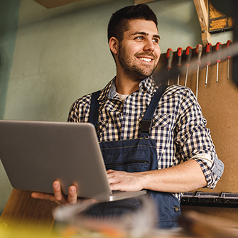 A smiling male tradesperson in a workshop using a laptop.