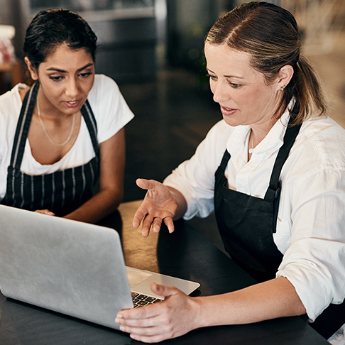 Two women in aprons look at a laptop screen.