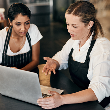 Two women in aprons look at a laptop screen.