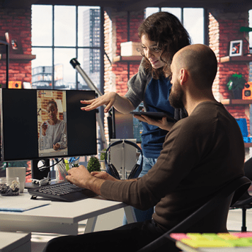 A man sits at a desk in front of a computer monitor as a woman gestures to the monitor.