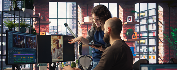 A man sits at a desk in front of a computer monitor as a woman gestures to the monitor.