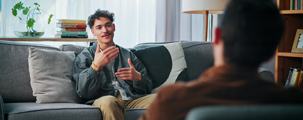 A man sits on a sofa with another person seated opposite him.