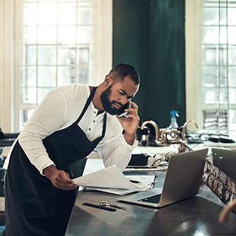 A man in an apron leans over a laptop while talking on the phone and holding a stack of papers.