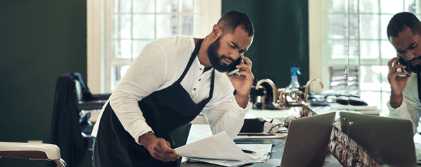 A man in an apron leans over a laptop while talking on the phone and holding a stack of papers.