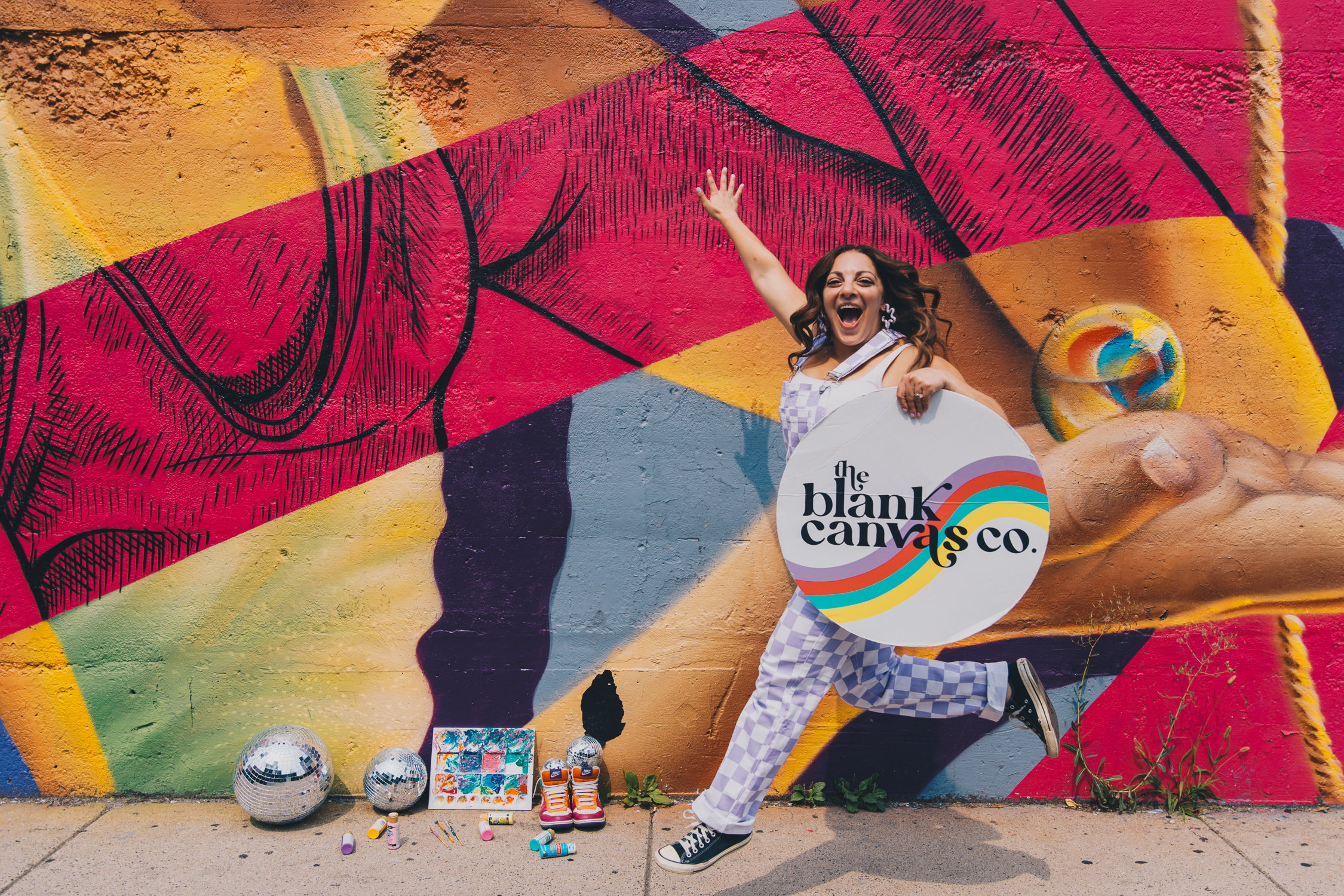 The Blank Canvas Co. founder Erica Meunier, jumping in front of a colorful mural holding a round sign that says The Blank Canvas Co.