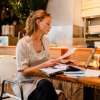 A woman in an apron sits at a restaurant table with a laptop adn paperwork.