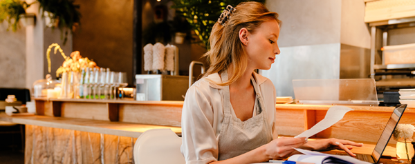 A woman in an apron sits at a restaurant table with a laptop adn paperwork.