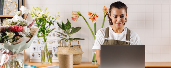 A florist works on her laptop surrounded by floral arrangements.