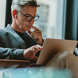 A man wearing earbuds sits in a chair looking down at his laptop.