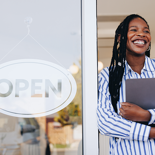 A woman holding a laptop to her chest stands in an open doorway next to an "Open" sign.