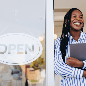 A woman holding a laptop to her chest stands in an open doorway next to an "Open" sign.