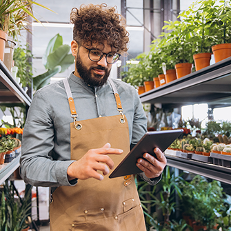 A small business owner wearing an apron and glasses standing in a plant nursery or flower shop while looking down at a digital tablet.