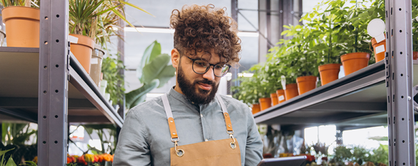 A small business owner wearing an apron and glasses standing in a plant nursery or flower shop while looking down at a digital tablet.
