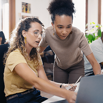 A woman sits at a desk in an office while another woman looks over her shoulder.