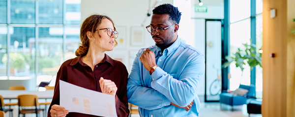 A man and a woman stand in an office looking down at a sheet of paper.