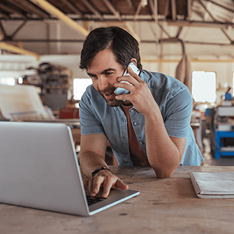A man leans on a desk in a workshop while talking on the phone and looking into a laptop screen.