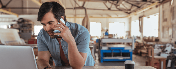 A man leans on a desk in a workshop while talking on the phone and looking into a laptop screen.