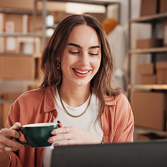 A woman holding a coffee cup sits at a desk looking into a laptop screen.