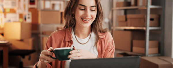 A woman holding a coffee cup sits at a desk looking into a laptop screen.