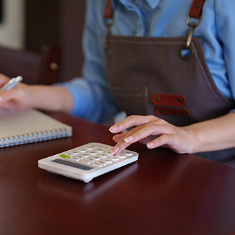A person wearing an apron uses a calculator with one hand and takes notes with the other.