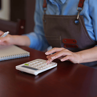 A person wearing an apron uses a calculator with one hand and takes notes with the other.