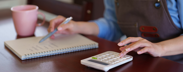 A person wearing an apron uses a calculator with one hand and takes notes with the other.
