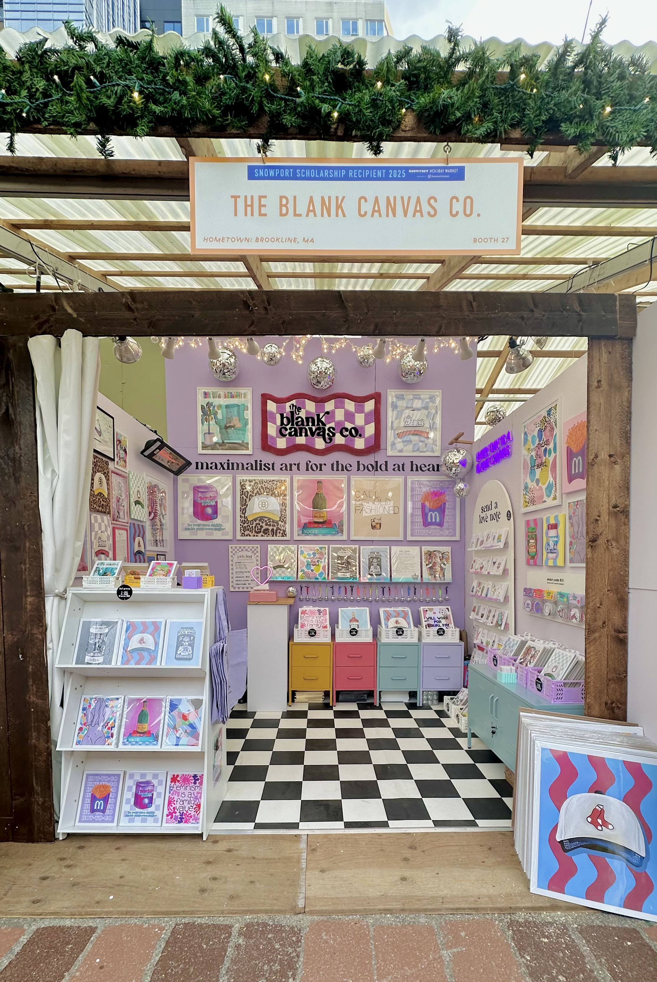 The Blank Canvas Co. colorful outdoor market booth with a black and white checkered floor, purple walls and artwork for sale. 