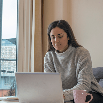A woman in a sweater sits in front of a window using a laptop.