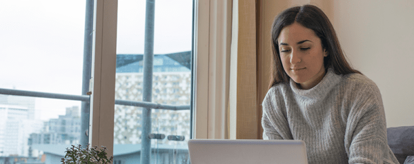 A woman in a sweater sits in front of a window using a laptop.