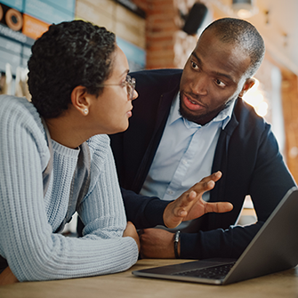 A woman and a man converse while huddled over a laptop in a cafe.
