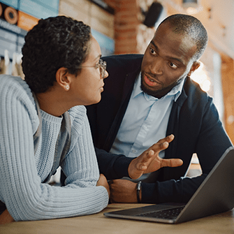A woman and a man converse while huddled over a laptop in a cafe.