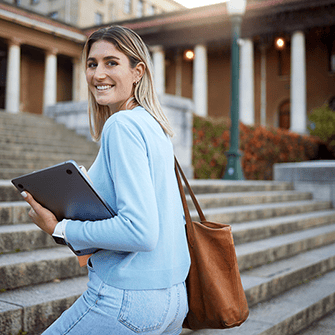 A woman stands on the steps of a university building carrying a tote bag and a laptop.