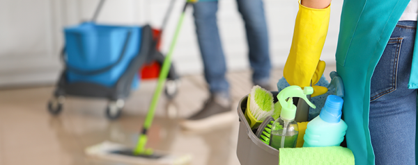 A person wearing yellow gloves carries a caddy of cleaning supplies.