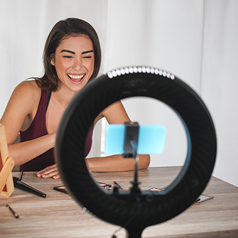 A woman smiles while filming herself on a smartphone positioned inside a ring light.