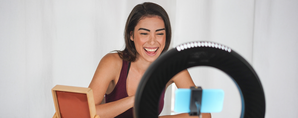 A woman smiles while filming herself on a smartphone positioned inside a ring light.