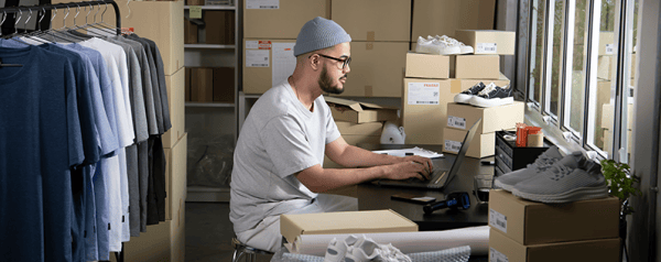 A man wearing a beanie and glasses works on a laptop in a warehouse room filled with stacked boxes, clothing racks, and shoes.