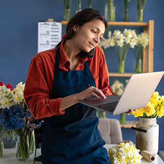 Florist wearing a blue apron uses a laptop in her flower shop, surrounded by colorful bouquets and floral arrangements.