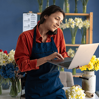 Florist wearing a blue apron uses a laptop in her flower shop, surrounded by colorful bouquets and floral arrangements.